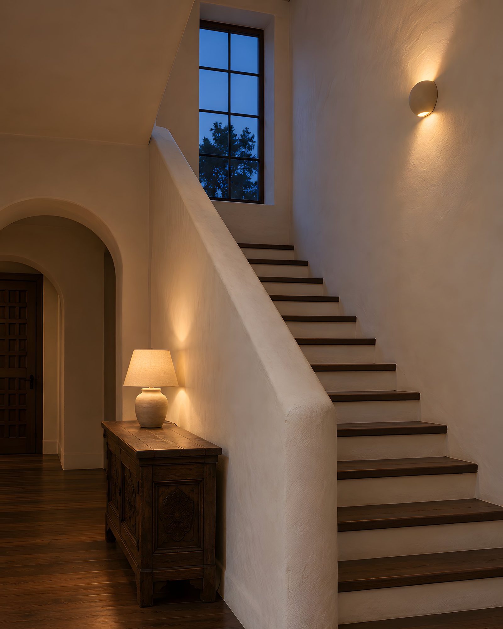 A plaster stair hall at dusk: stairs rising along a curved wall, a glowing wall lamp, a deep blue window above, a table lamp on a dark chest.